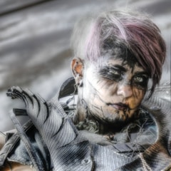 A person with short, dyed hair styled in a futuristic or post-apocalyptic fashion. The face is elaborately painted with dark makeup, creating an intense and artistic look. The attire includes textured, armor-like material with intricate details, contributing to a bold and edgy aesthetic.