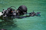 A scuba diver is floating in clear turquoise water. The diver is wearing a black wetsuit and is equipped with a large camera housing, possibly for underwater photography. The sunlight creates a glistening effect on the surface of the water and on the diver's gear.
