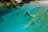 A diver wearing scuba gear floats in a clear, turquoise water body surrounded by lush greenery. The sunlight filters through the branches, casting shadows on the water. Stones and pebbles are visible at the shallow edges.
