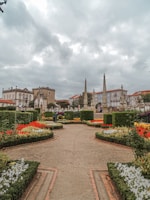 A beautifully landscaped garden features vibrant flowerbeds with a variety of colorful flowers arranged in geometric patterns. Pathways lead through the garden, bordered by manicured hedges. Historical buildings with intricate architecture and tall stone pillars can be seen in the background under a cloudy sky.