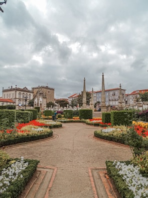 A beautifully landscaped garden features vibrant flowerbeds with a variety of colorful flowers arranged in geometric patterns. Pathways lead through the garden, bordered by manicured hedges. Historical buildings with intricate architecture and tall stone pillars can be seen in the background under a cloudy sky.