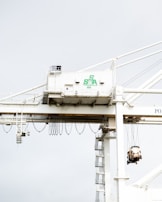 A large industrial crane is situated in a port setting. The crane has a white structure with a green logo that reads 'SSA Terminals.' The crane features various cables and machinery, including a suspended container or object being lifted. The background is a pale sky, creating a stark contrast with the crane's structure.