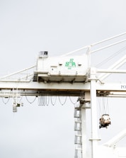 A large industrial crane is situated in a port setting. The crane has a white structure with a green logo that reads 'SSA Terminals.' The crane features various cables and machinery, including a suspended container or object being lifted. The background is a pale sky, creating a stark contrast with the crane's structure.