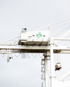 A large industrial crane is situated in a port setting. The crane has a white structure with a green logo that reads 'SSA Terminals.' The crane features various cables and machinery, including a suspended container or object being lifted. The background is a pale sky, creating a stark contrast with the crane's structure.