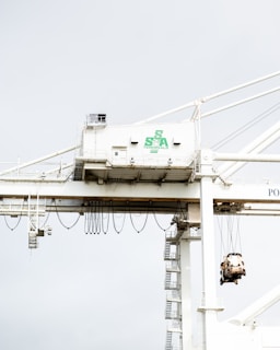 A large industrial crane is situated in a port setting. The crane has a white structure with a green logo that reads 'SSA Terminals.' The crane features various cables and machinery, including a suspended container or object being lifted. The background is a pale sky, creating a stark contrast with the crane's structure.