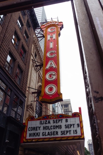 A large vertical marquee sign with the word 'CHICAGO' in bold letters hangs from a classic theater building. The sign is red with white letters and is bordered with lights. Below, there is a board listing upcoming events and dates, including shows by Iliza, Corey Holcomb, and Nikki Glaser. Surrounding the marquee are historic brick and stone buildings, hinting at an urban setting.