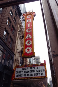 A large vertical marquee sign with the word 'CHICAGO' in bold letters hangs from a classic theater building. The sign is red with white letters and is bordered with lights. Below, there is a board listing upcoming events and dates, including shows by Iliza, Corey Holcomb, and Nikki Glaser. Surrounding the marquee are historic brick and stone buildings, hinting at an urban setting.