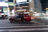 A red taxi with the branding 'S.RIDE' is moving quickly across a city street at night. The vehicle is captured in motion, creating a dynamic blur effect against a backdrop of brightly lit buildings and neon signs. The scene conveys a sense of urban hustle and vibrant nightlife.