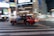 A red taxi with the branding 'S.RIDE' is moving quickly across a city street at night. The vehicle is captured in motion, creating a dynamic blur effect against a backdrop of brightly lit buildings and neon signs. The scene conveys a sense of urban hustle and vibrant nightlife.