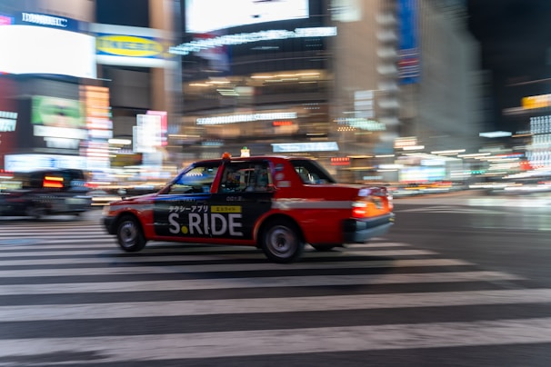A red taxi with the branding 'S.RIDE' is moving quickly across a city street at night. The vehicle is captured in motion, creating a dynamic blur effect against a backdrop of brightly lit buildings and neon signs. The scene conveys a sense of urban hustle and vibrant nightlife.