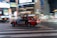 A red taxi with the branding 'S.RIDE' is moving quickly across a city street at night. The vehicle is captured in motion, creating a dynamic blur effect against a backdrop of brightly lit buildings and neon signs. The scene conveys a sense of urban hustle and vibrant nightlife.