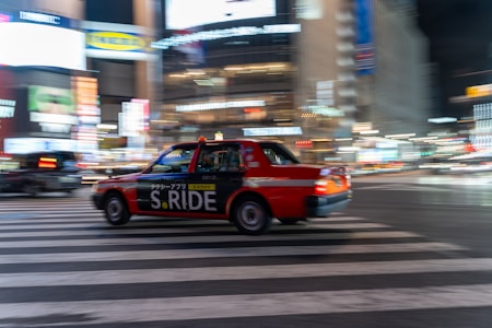 A red taxi with the branding 'S.RIDE' is moving quickly across a city street at night. The vehicle is captured in motion, creating a dynamic blur effect against a backdrop of brightly lit buildings and neon signs. The scene conveys a sense of urban hustle and vibrant nightlife.