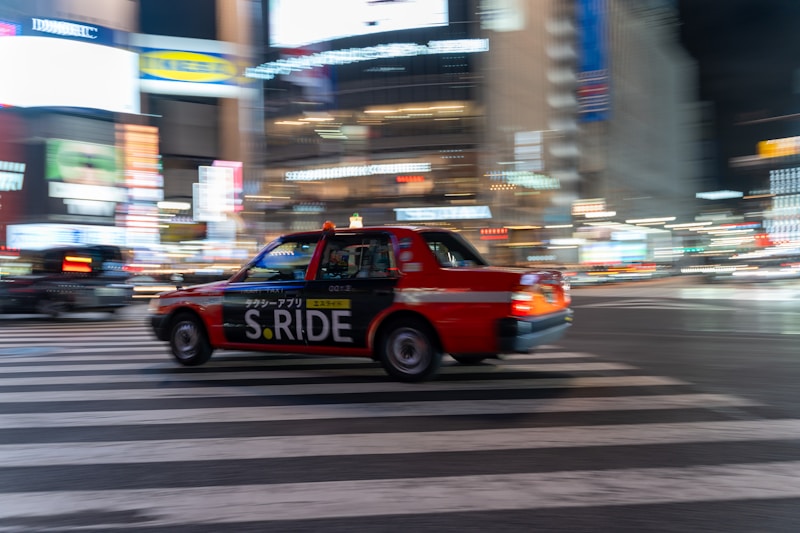 A red taxi with the branding 'S.RIDE' is moving quickly across a city street at night. The vehicle is captured in motion, creating a dynamic blur effect against a backdrop of brightly lit buildings and neon signs. The scene conveys a sense of urban hustle and vibrant nightlife.