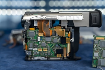 A partially disassembled digital video camera with exposed electronic components. The camera's buttons for focus, fade, white balance, and backlight compensation are visible. The green circuit boards with various chips, connectors, and ribbon cables are prominently displayed, with additional circuit boards in the background.