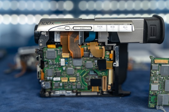 A partially disassembled digital video camera with exposed electronic components. The camera's buttons for focus, fade, white balance, and backlight compensation are visible. The green circuit boards with various chips, connectors, and ribbon cables are prominently displayed, with additional circuit boards in the background.