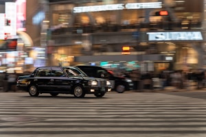 A black taxi moves across a busy urban street at night, surrounded by blurred lights and bustling activity. The background features brightly lit signs and glass-fronted buildings, indicating a vibrant city atmosphere.