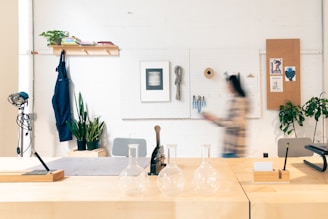 A well-organized workspace featuring a wooden table with three glass flasks, a pencil, and notepad. The background shows a pegboard with hanging tools and artwork. A blurred person walks past, adding movement to the scene. Several plants and a blue apron hang from a shelf, alongside books adding a cozy, creative touch.