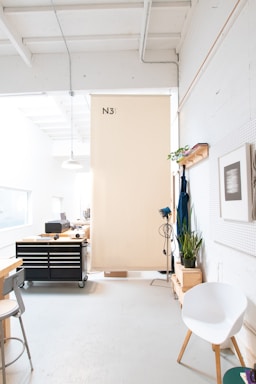 A well-lit, modern workspace with high ceilings and white walls. There is a large wooden board or divider with the label 'N3' hanging from the ceiling. On the left side, there's a black rolling tool chest with various items on top, and a tall stool next to a countertop. Towards the back, there is office equipment, including a printer. The right side features a white chair, a potted plant, an apron hanging on the wall, and a framed picture. Above the picture is a shelf with a small plant and a book.