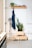 A minimalistic interior features a white wall with a wooden shelf holding books, a small potted plant, and decorative items. Below, a wooden peg holds a dark blue apron, and two potted plants sit atop a stack of wooden crates. On the left, a tall, vintage-style lamp stands next to a wooden storage unit.