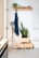 A minimalistic interior features a white wall with a wooden shelf holding books, a small potted plant, and decorative items. Below, a wooden peg holds a dark blue apron, and two potted plants sit atop a stack of wooden crates. On the left, a tall, vintage-style lamp stands next to a wooden storage unit.