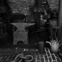 An old workshop setting with tools and equipment related to blacksmithing. A large anvil sits in the center, secured with chains. The background features a rustic brick wall and various metalworking tools, including a watering can and heavy chains. The atmosphere is industrial and nostalgic, suggesting a historical or traditional working environment.