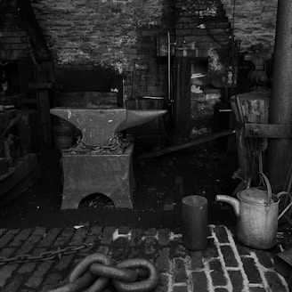 An old workshop setting with tools and equipment related to blacksmithing. A large anvil sits in the center, secured with chains. The background features a rustic brick wall and various metalworking tools, including a watering can and heavy chains. The atmosphere is industrial and nostalgic, suggesting a historical or traditional working environment.