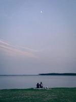 A couple sitting comfortably on a moisture-proof mat beside a tranquil lake at sunset.