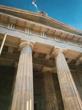 A neoclassical building with tall, impressive columns extends upwards towards a vibrant blue sky. The Greek flag flutters at the top of the structure, and intricate stone carvings adorn the façade.