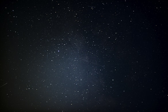 A night sky filled with numerous stars scattered across a dark background. A subtle, glowing band crosses the center, suggesting the Milky Way. A faint meteor streak is visible, adding a dynamic element to the stillness.