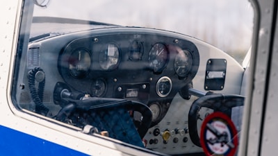 An airplane cockpit interior with a range of analog dials and gauges on the instrument panel, a steering yoke prominently in the foreground, and a two-way radio microphone attached on the left.