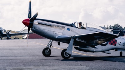 A vintage military aircraft is parked on an airfield. A pilot wearing a helmet and goggles is seated in the open cockpit. The plane features a polished metal body with distinctive checkered and roundel markings on the fuselage. In the background, there are additional aircraft and hangars under a partly cloudy sky.
