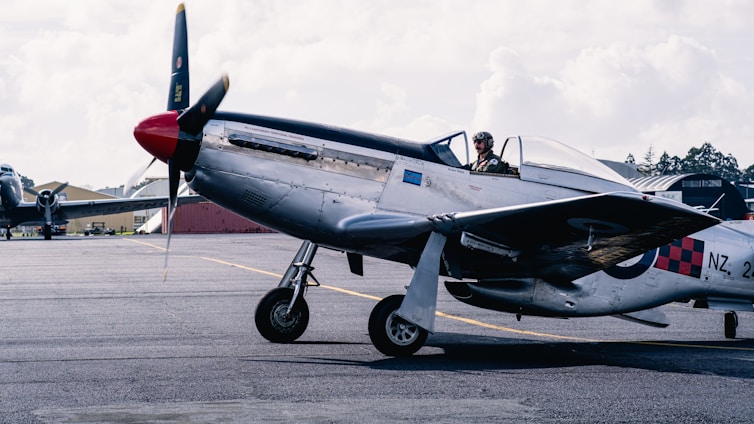 A pilot wearing a check six co. jacket standing beside a vintage airplane on a sunny airfield.