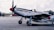 A vintage military aircraft is parked on an airfield. A pilot wearing a helmet and goggles is seated in the open cockpit. The plane features a polished metal body with distinctive checkered and roundel markings on the fuselage. In the background, there are additional aircraft and hangars under a partly cloudy sky.