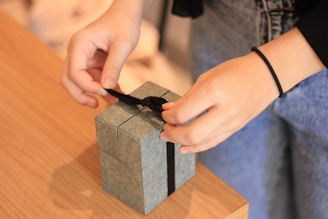 Hands carefully tying a black ribbon on a small gray gift box placed on a wooden table. The person is wearing casual clothing, including a black sleeve and blue jeans.