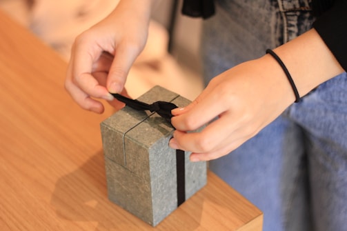 Hands carefully tying a black ribbon on a small gray gift box placed on a wooden table. The person is wearing casual clothing, including a black sleeve and blue jeans.