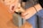 Hands carefully tying a black ribbon on a small gray gift box placed on a wooden table. The person is wearing casual clothing, including a black sleeve and blue jeans.