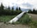 A large metal pipeline extends through a forested area, surrounded by dense greenery and trees. The sky is overcast, suggesting the possibility of rain. The pipeline runs parallel to a gravel road, indicating an infrastructure passing through a natural environment.