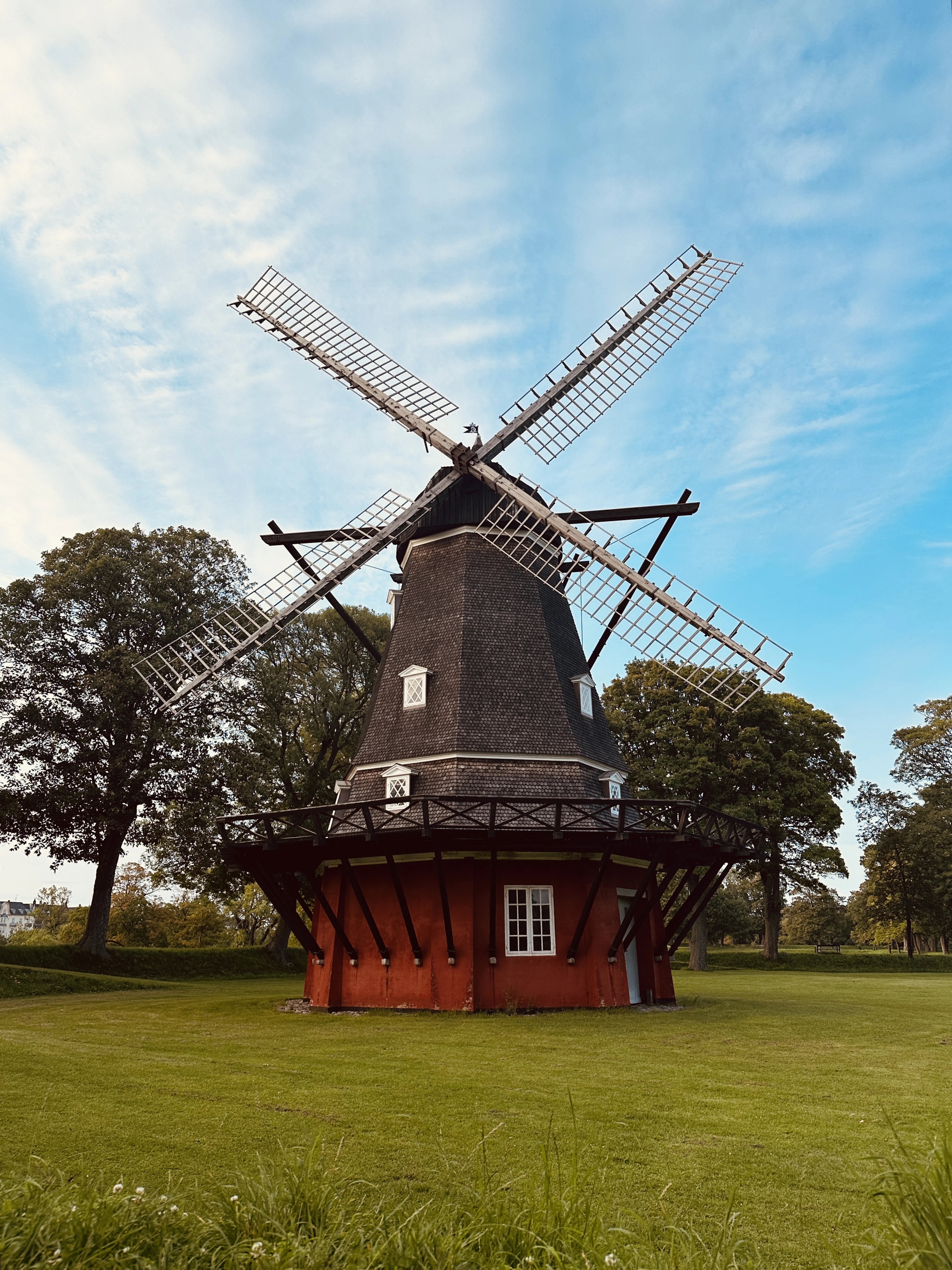 The red-timbered windmill standing on the grass ramparts of Kastellet in Copenhagen, its white sails spread wide against a blue sky streaked with high cloud.