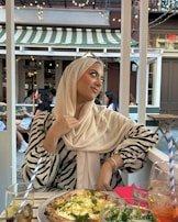 A person wearing a beige hijab and zebra-striped top sits at an outdoor café. She is looking to the side with a pleasant expression, holding the edge of her hijab. On the table in front of her is a pizza and glasses with beverages. The background shows other people dining and string lights hanging from above, under a green and white striped awning.