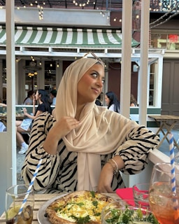 A person wearing a beige hijab and zebra-striped top sits at an outdoor café. She is looking to the side with a pleasant expression, holding the edge of her hijab. On the table in front of her is a pizza and glasses with beverages. The background shows other people dining and string lights hanging from above, under a green and white striped awning.