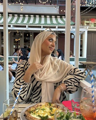 A person wearing a beige hijab and zebra-striped top sits at an outdoor café. She is looking to the side with a pleasant expression, holding the edge of her hijab. On the table in front of her is a pizza and glasses with beverages. The background shows other people dining and string lights hanging from above, under a green and white striped awning.
