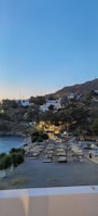 A serene coastal scene at dusk, featuring a beach with organized rows of sunbeds and umbrellas. The landscape in the background is hilly with scattered white houses and lush greenery. The area is illuminated by warm lights, creating a cozy atmosphere.