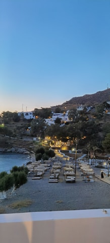 A serene coastal scene at dusk, featuring a beach with organized rows of sunbeds and umbrellas. The landscape in the background is hilly with scattered white houses and lush greenery. The area is illuminated by warm lights, creating a cozy atmosphere.