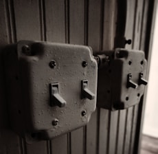 Two vintage, metal toggle light switches mounted on a textured, vertically paneled wooden wall. The switches appear worn, with visible screws and a slightly rusted finish, giving an aged industrial feel.