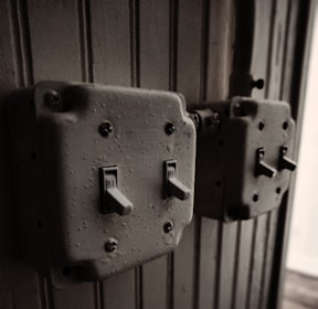 Two vintage, metal toggle light switches mounted on a textured, vertically paneled wooden wall. The switches appear worn, with visible screws and a slightly rusted finish, giving an aged industrial feel.