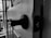A close-up view of a door ajar with a door latch and handle visible in the foreground, casting shadows. The background shows a glimpse of the outside through a window.