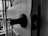 A close-up view of a door ajar with a door latch and handle visible in the foreground, casting shadows. The background shows a glimpse of the outside through a window.