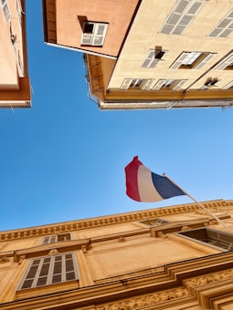 A French flag flutters atop a beige building with detailed architectural features. Two adjacent buildings frame a clear blue sky.