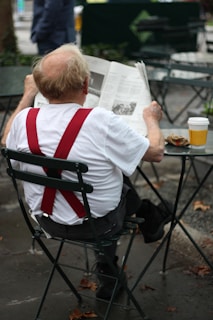 An elderly person with red suspenders is sitting on a metal chair outdoors, reading a newspaper. A takeaway coffee cup is placed on a table next to them. The setting appears to be a park or an outdoor café, with fallen leaves scattered on the ground.
