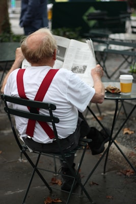 An elderly person with red suspenders is sitting on a metal chair outdoors, reading a newspaper. A takeaway coffee cup is placed on a table next to them. The setting appears to be a park or an outdoor café, with fallen leaves scattered on the ground.